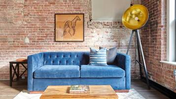 Une pièce avec un canapé bleu, une table basse en bois, un tableau encadré représentant un cheval, un lampadaire et un mur en briques.