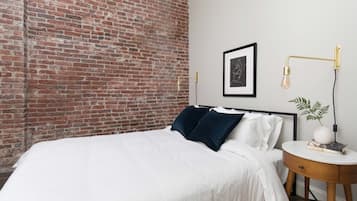 Une chambre avec un mur en briques, un lit avec une literie blanche, une table de chevet avec une lampe et un tableau encadré accroché au mur.