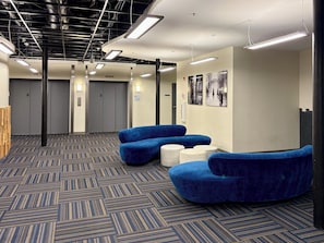 A waiting area with blue seating, a patterned carpet, and a ceiling with exposed pipes. - Kasa At Boston Common (Boston)
