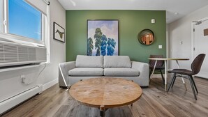 Une pièce comprenant une table ronde en bois, un canapé gris, une table à manger avec des chaises, une fenêtre avec des stores et un mur vert orné d’un tableau encadré.