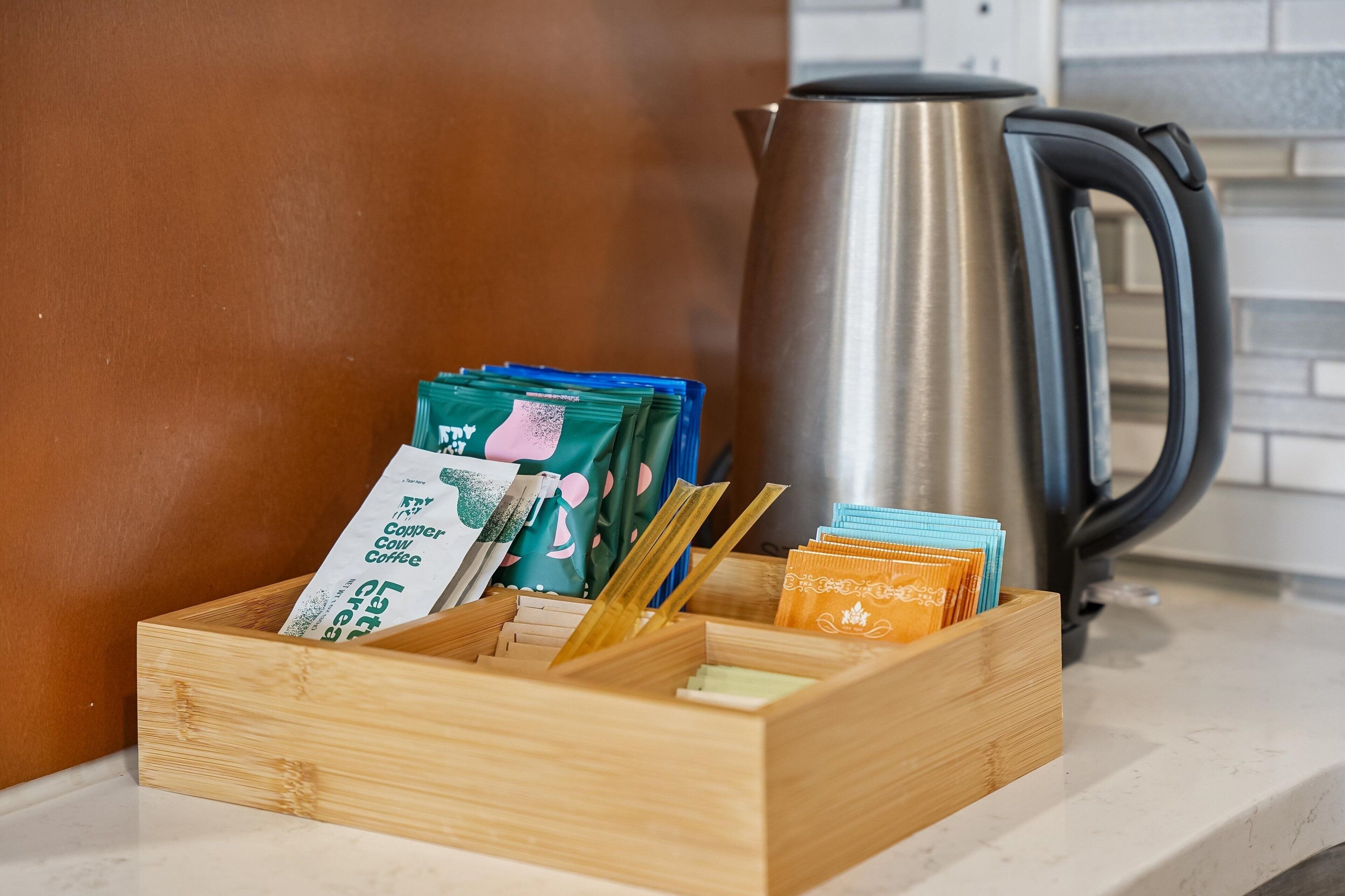 A wooden box with coffee packets and a stainless steel kettle on a kitchen counter.