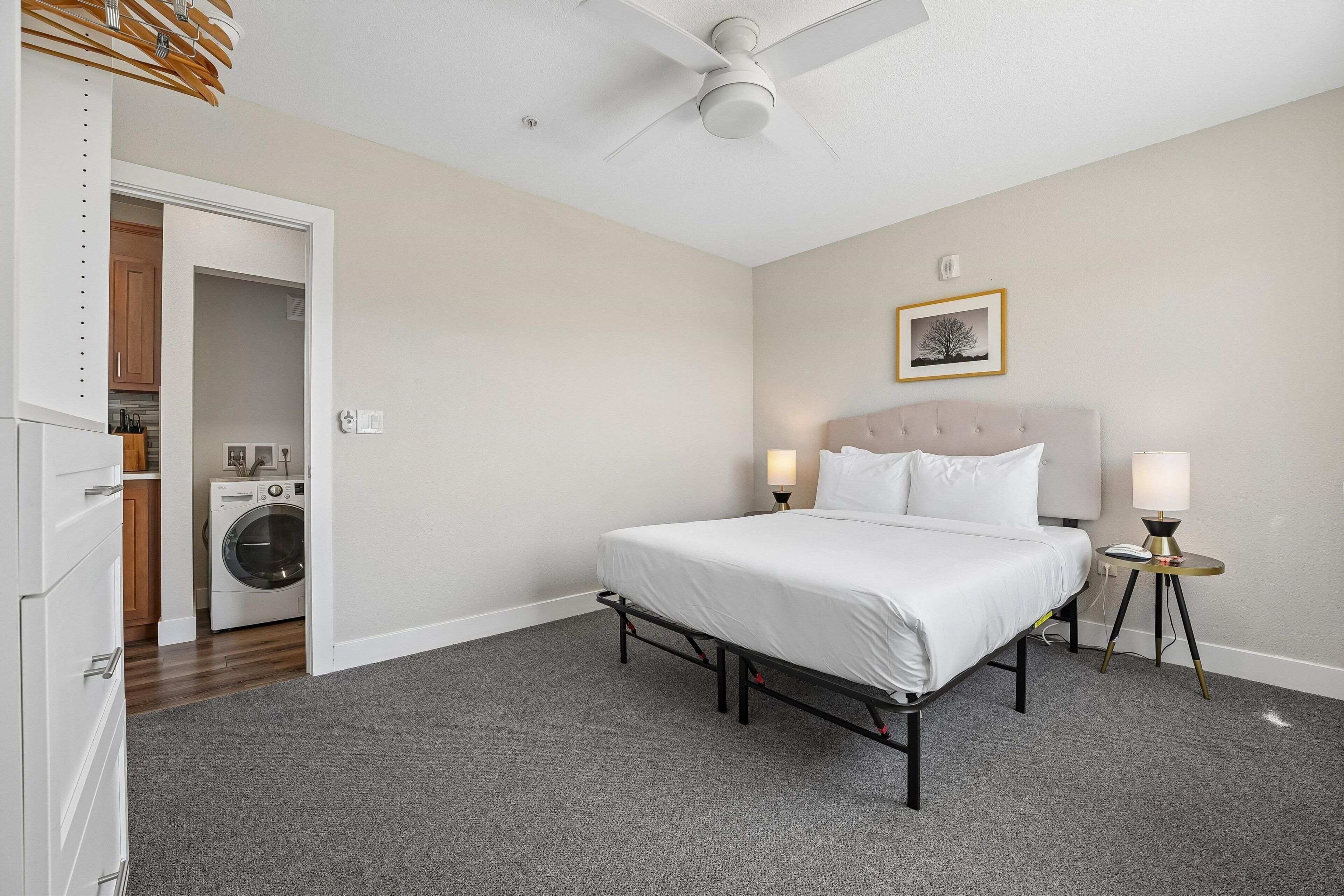 Ein Schlafzimmer mit Bett, Nachttischlampen, Deckenventilator, einer Waschmaschine und einer Tür zu einer Küche.