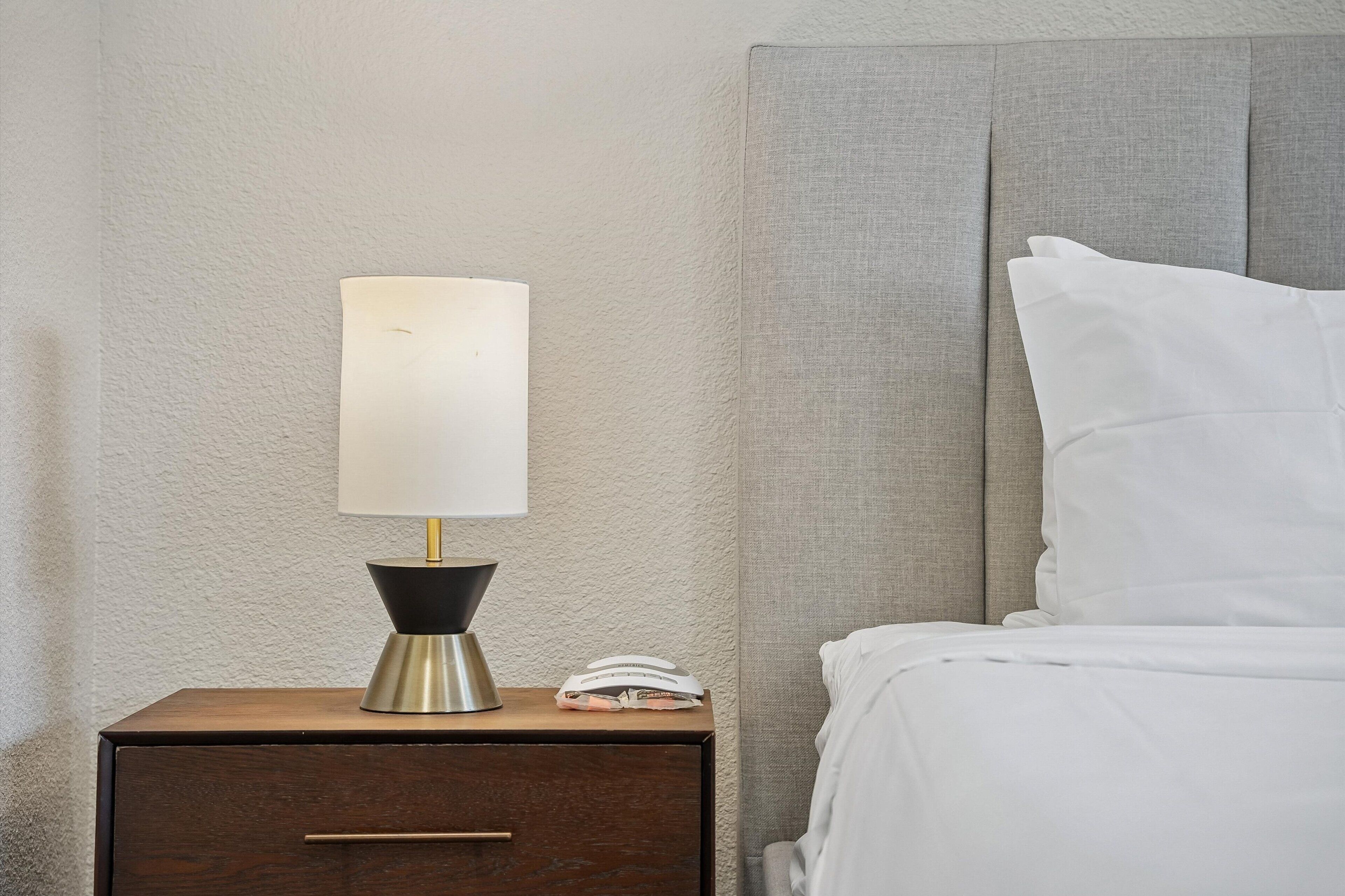 Un lit bien fait, avec du linge de lit blanc, une lampe de chevet à abat-jour blanc et un livre fermé posé sur une table de chevet en bois.