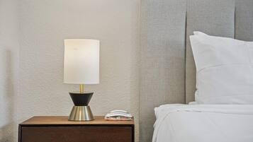 Un lit bien fait, avec du linge de lit blanc, une lampe de chevet à abat-jour blanc et un livre fermé posé sur une table de chevet en bois.