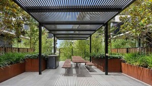 Un espace extérieur abrité, doté d’une table et de bancs, entouré de jardinières et d’une clôture en métal.