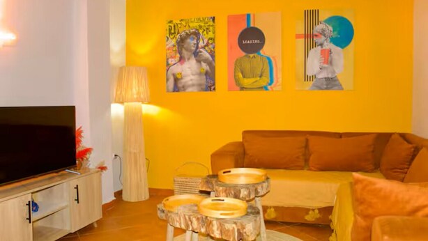 Ein Wohnzimmer mit Fernseher, Couchtisch aus Holz, orangefarbenem Sofa und Wandbild.