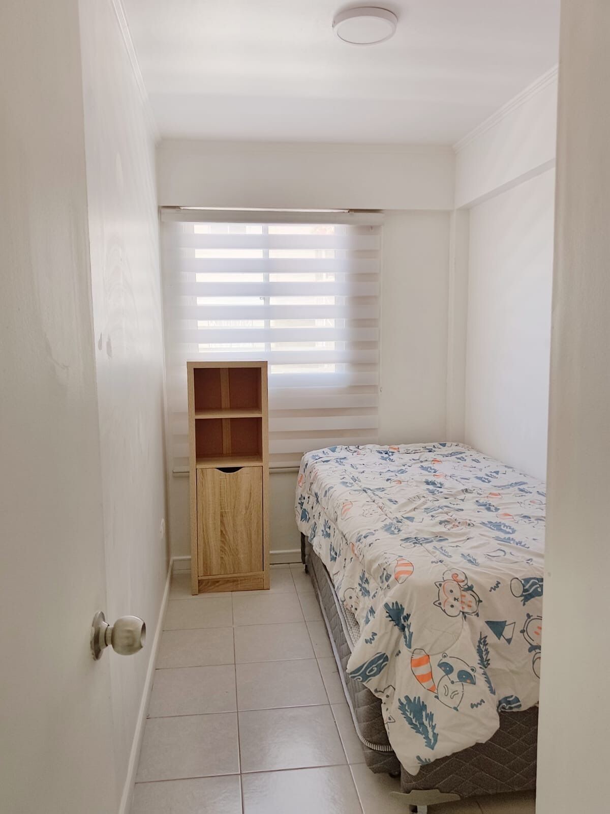 A small, clean room with a bed, a wooden cabinet, and a window with blinds.
