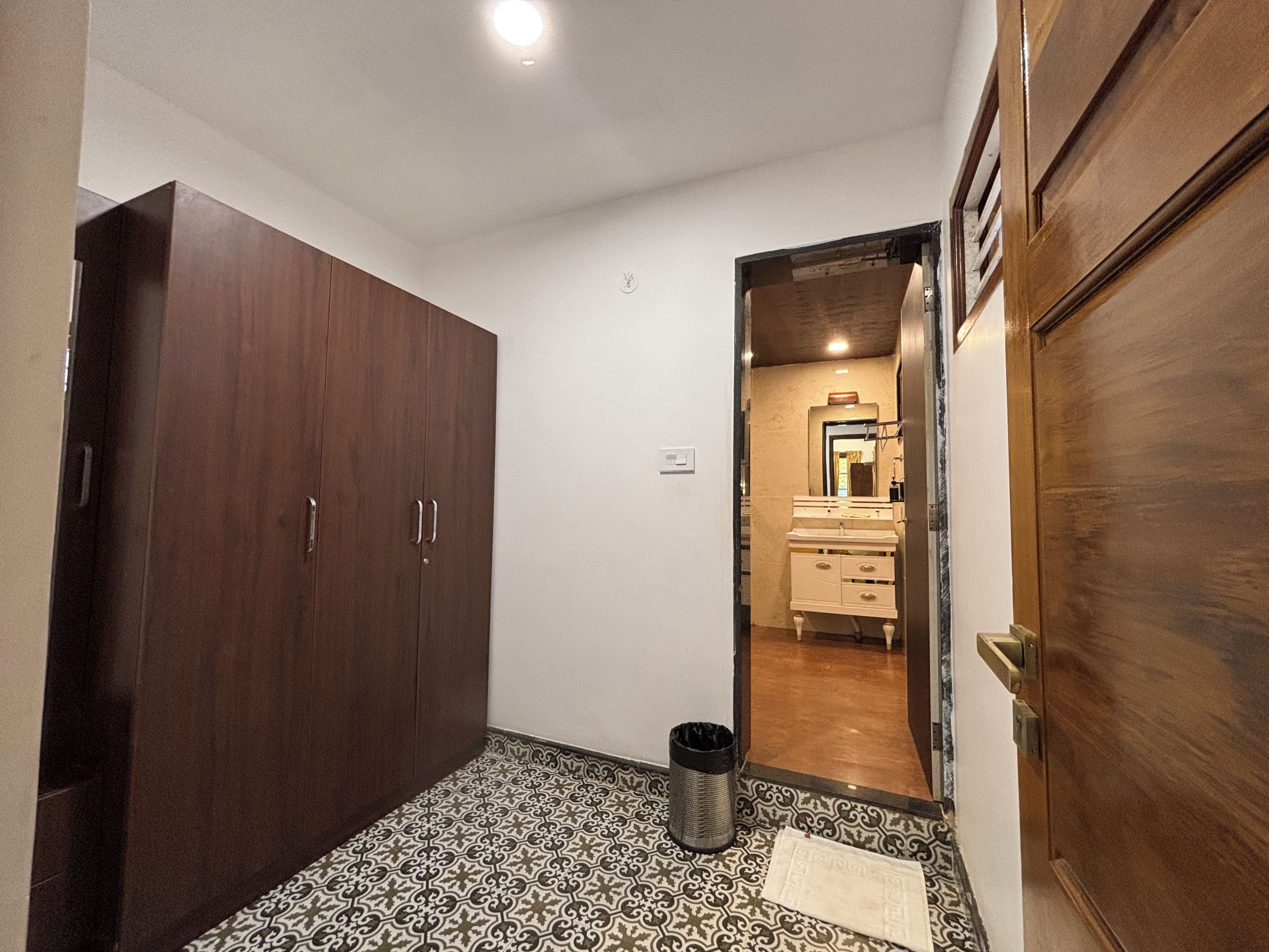 A hallway with a wardrobe, a trash bin, patterned floor tiles, and a door leading to a bathroom with a mirror and sink.