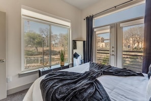 A bedroom with a large window, a bed, a nightstand, and a view of trees and buildings. - Riverwalk Oasis - Pool, Free Parking & Views (San Antonio)