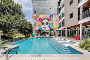 A swimming pool with a mural on the adjacent building, surrounded by greenery and modern architecture. - Riverwalk Oasis - Pool, Free Parking & Views (San Antonio)