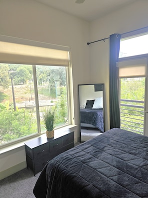 Een slaapkamer met een groot raam, een bed, een dressoir en een spiegel.
