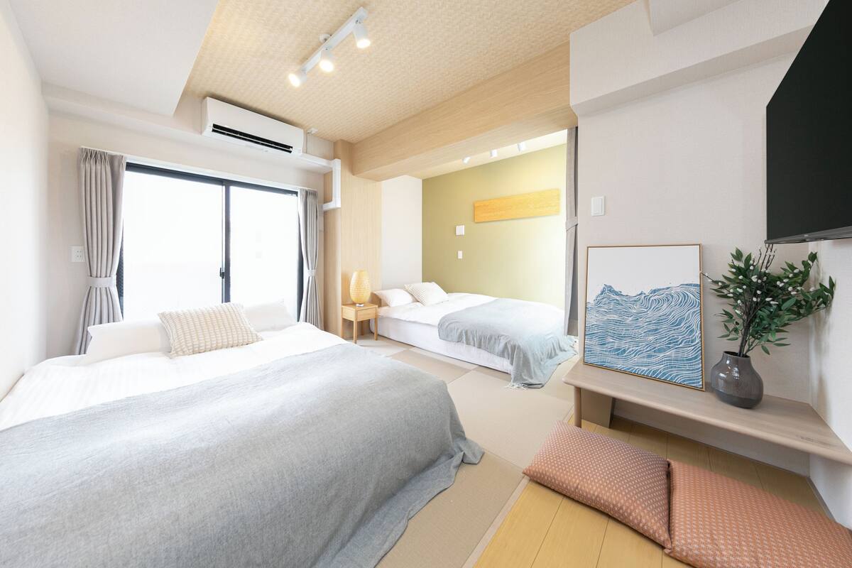 A hotel room with a large bed, a smaller bed, a bedside table with a lamp, a wall-mounted TV, and a painting.