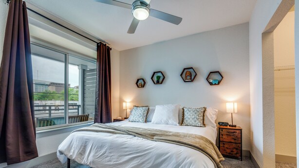 Quarto com cama grande, ventoinha no teto, janela com cortinas, luminárias de cabeceira e arte decorativa na parede.