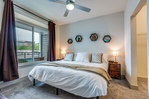 Un dormitorio con una cama grande, un ventilador de techo, una ventana con cortinas, lámparas de noche y decoración en la pared.