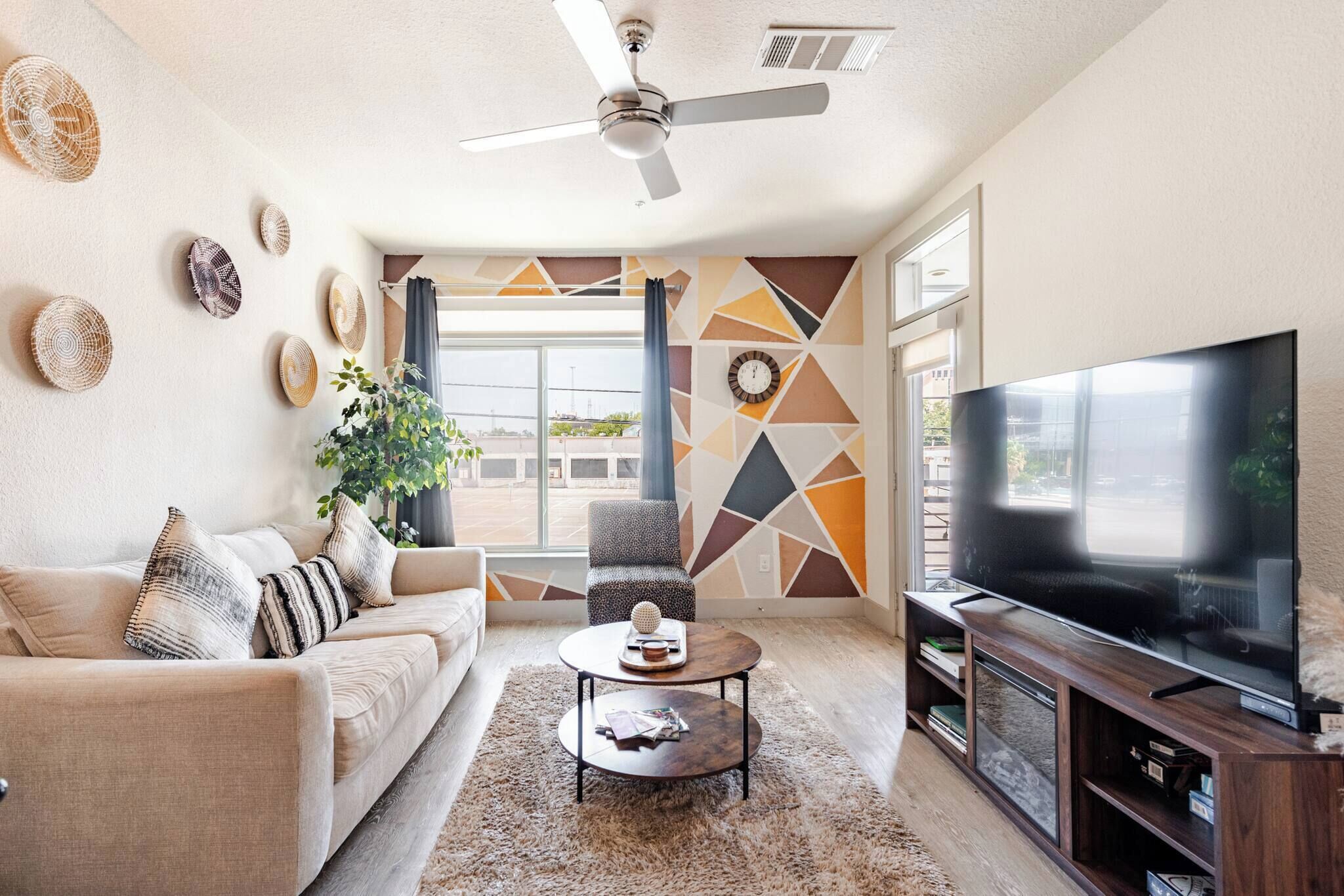 Sala de estar con sofá, mesa de centro, televisor, ventilador de techo, decoración en la pared y un ventanal grande.