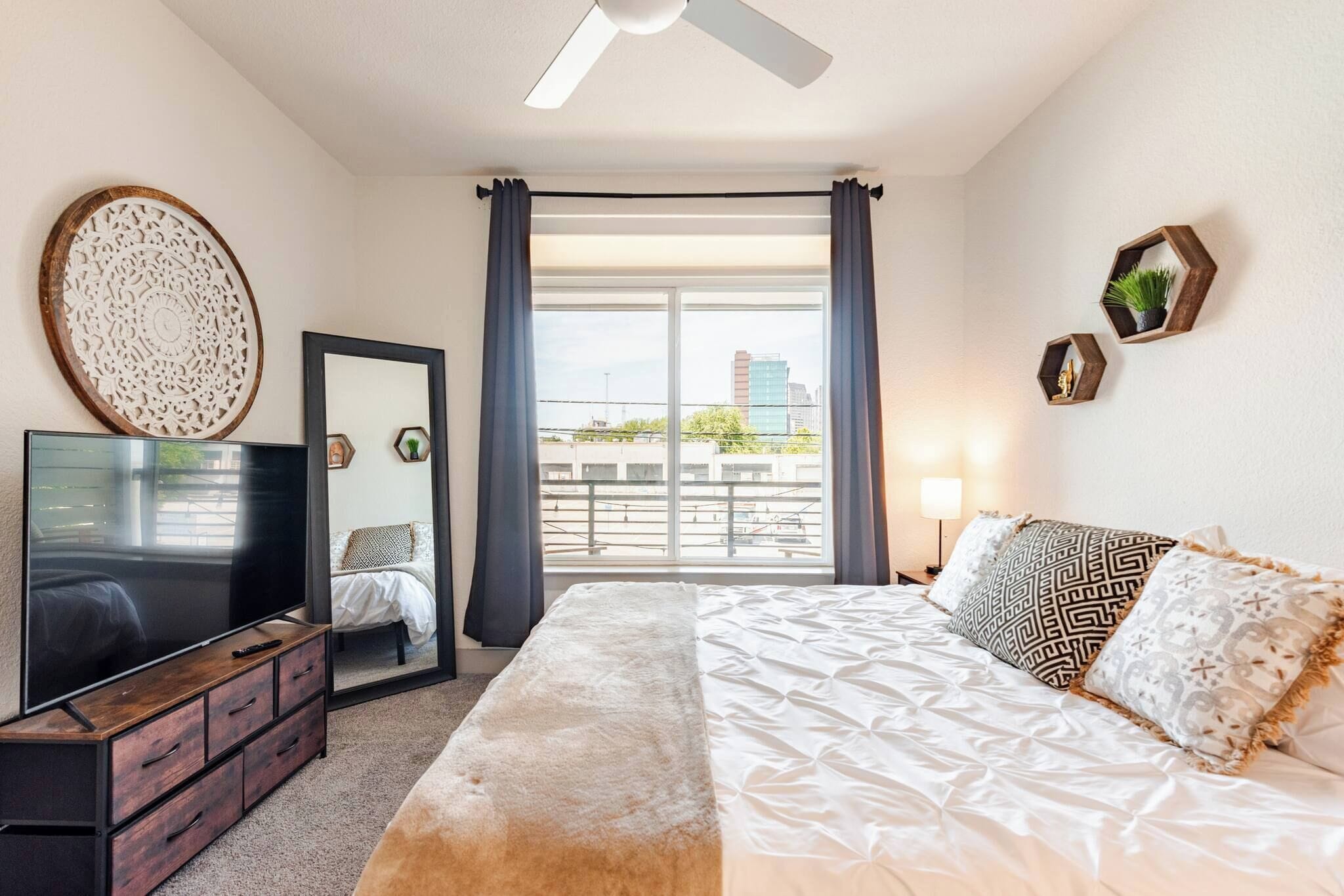 Un dormitorio con cama, televisión, cómoda, espejo y vista al balcón.