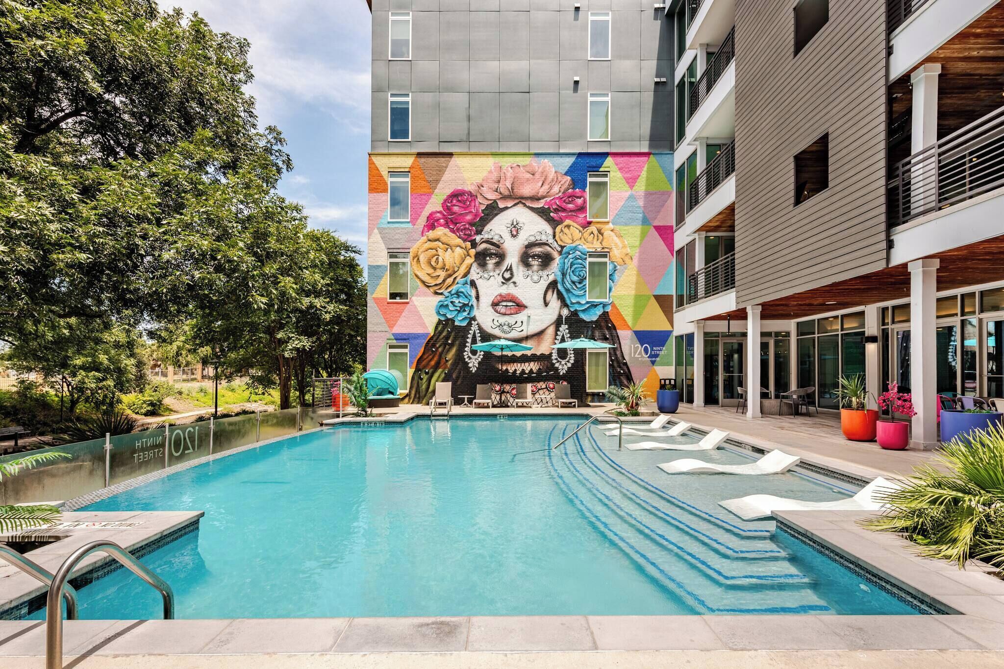 Una piscina con un mural, rodeada por un edificio moderno con balcones y una zona ajardinada.