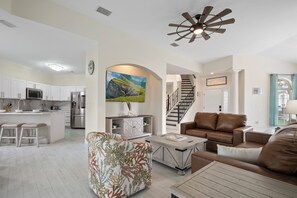 A modern kitchen with white cabinets, a central island, and stainless steel appliances. There is a living area with a brown leather sofa, a patterned armchair, and a coffee table. A staircase leads to an upper level. - SOUTH-FACING POOL, SPA, THEATER, GAMEROOM NEAR DISNEY (Haines City)