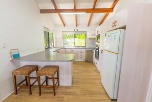 A kitchen with a central island, wooden stools, a refrigerator, and a window with a view of greenery. - Amity Treetops - beautiful coastal escape (Amity)