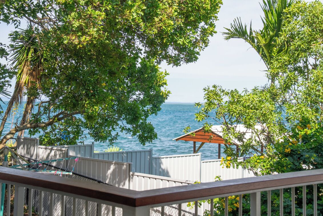 A view of the ocean from a balcony with a wooden railing, surrounded by lush green foliage and a small wooden structure.