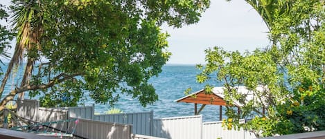 A view of the ocean from a balcony with a wooden railing, surrounded by lush green foliage and a small wooden structure.