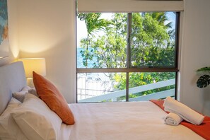 A bedroom with a bed, pillows, a nightstand, a lamp, a window with a view of trees and a balcony. - Amity Treetops - beautiful coastal escape (Amity)
