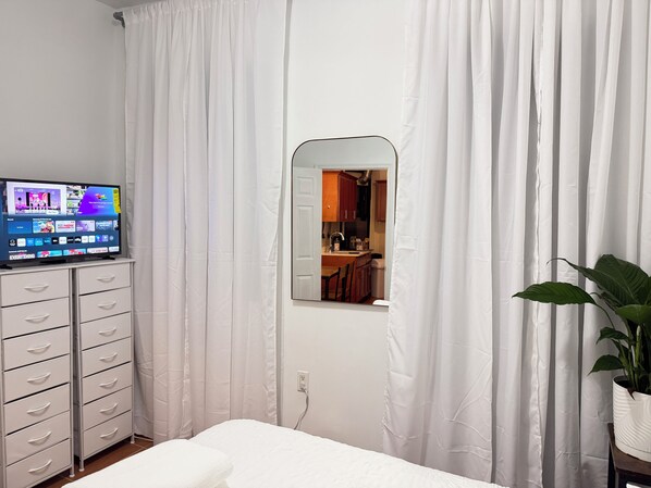 A bedroom with a TV, a wardrobe, a mirror, a window, and a potted plant.