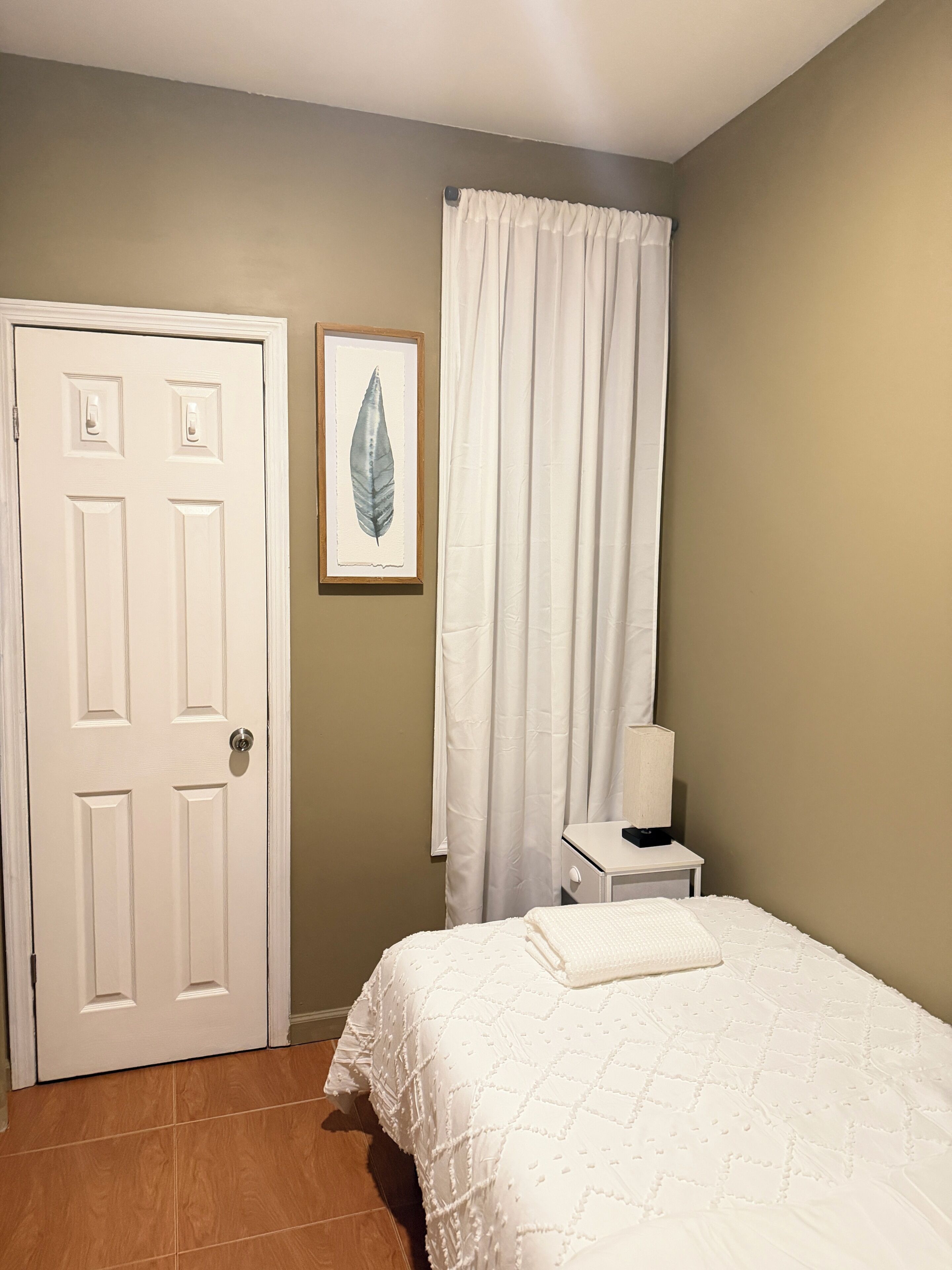 A bedroom with a white door, a bed with a white quilt, a nightstand with a lamp, and a curtain.