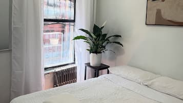 Een slaapkamer met een bed, een radiator, een raam met gordijnen, een kamerplant en een schilderij aan de muur.