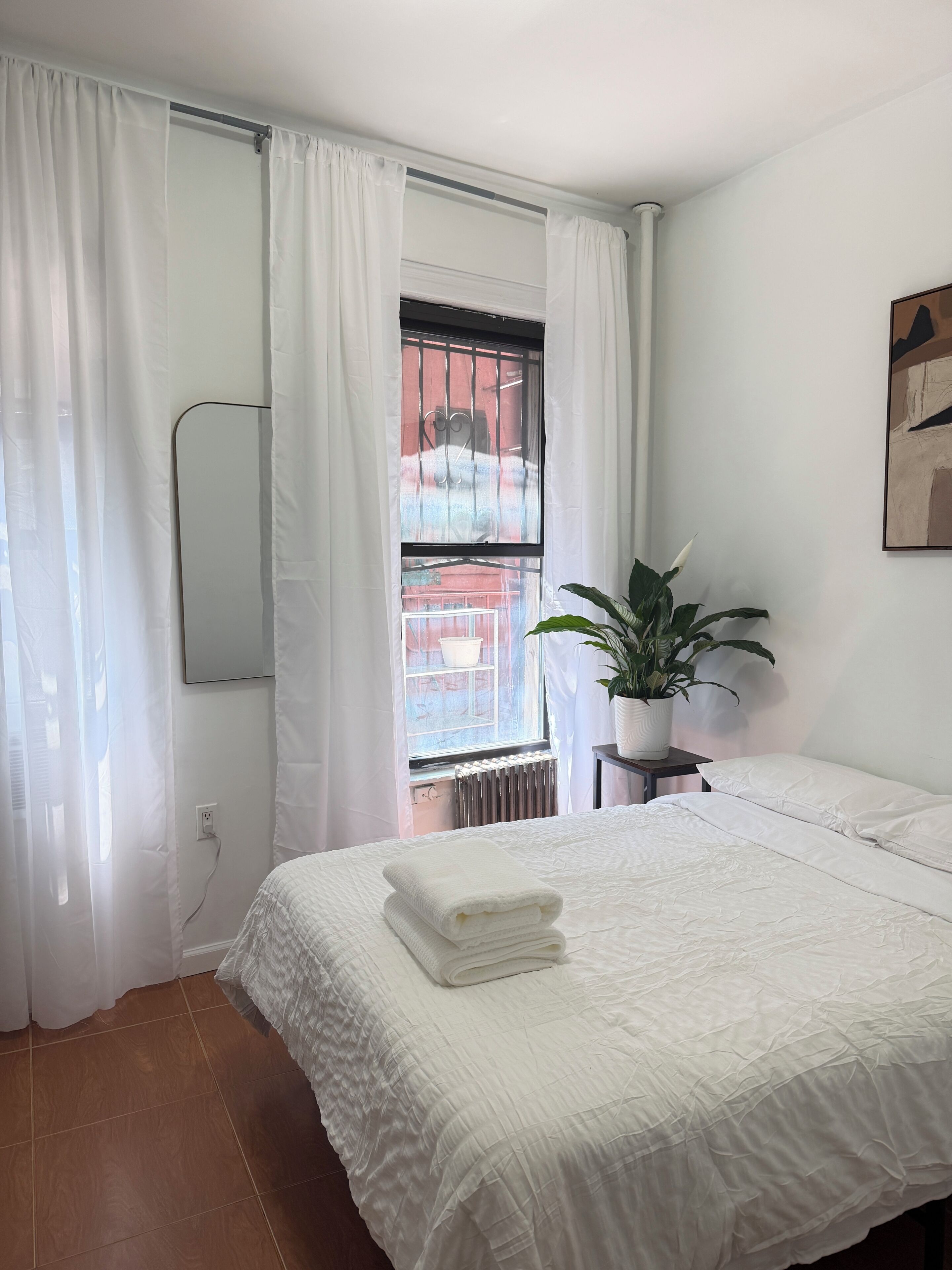 Een slaapkamer met een bed, witte gordijnen, een potplant en een radiator.