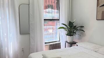 Een slaapkamer met een bed, witte gordijnen, een potplant en een radiator.