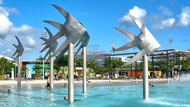 Een zwembad met grote metalen sculpturen en een gebouw met een groen-blauwe gevel op de achtergrond.