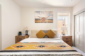 Une chambre à coucher avec un lit, une table de chevet, une lampe, une horloge et une fenêtre donnant sur des immeubles.