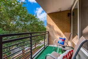 Un balcón con vistas a árboles y un muro de ladrillo, una silla con un cojín de Texas, y una mesita.