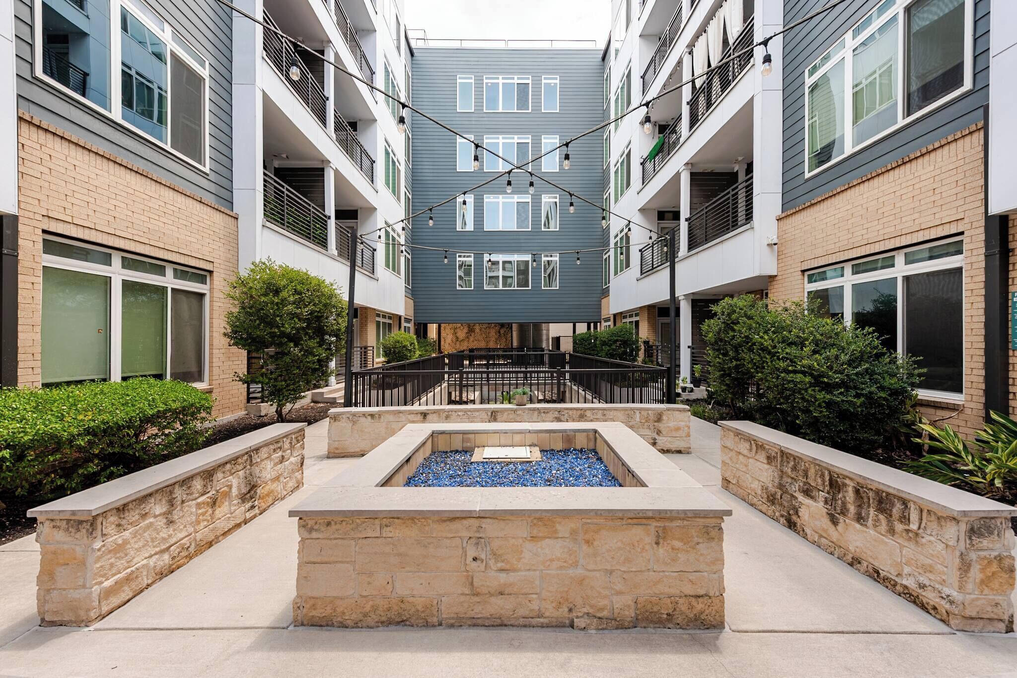 Un immeuble résidentiel à plusieurs étages, doté de balcons, d’une cour intérieure agrémentée d’un élément aquatique et d’un mur de soutènement en pierre.