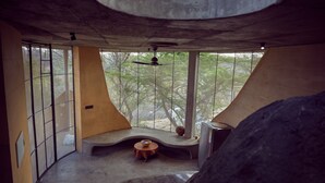 A room with large windows, a ceiling fan, a small table with a bowl, and a stone-like structure. - Winterfell by Sanctity Ferme Stays (Shoolagiri)