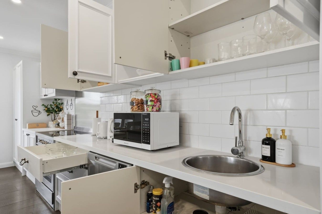 A modern kitchen with white cabinetry, a built-in microwave, a stainless steel sink, and a shelf with glassware and bottles.