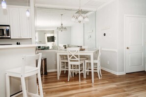 Una cocina moderna con armarios blancos, un microondas de acero inoxidable y una zona de comedor con sillas blancas y una araña.