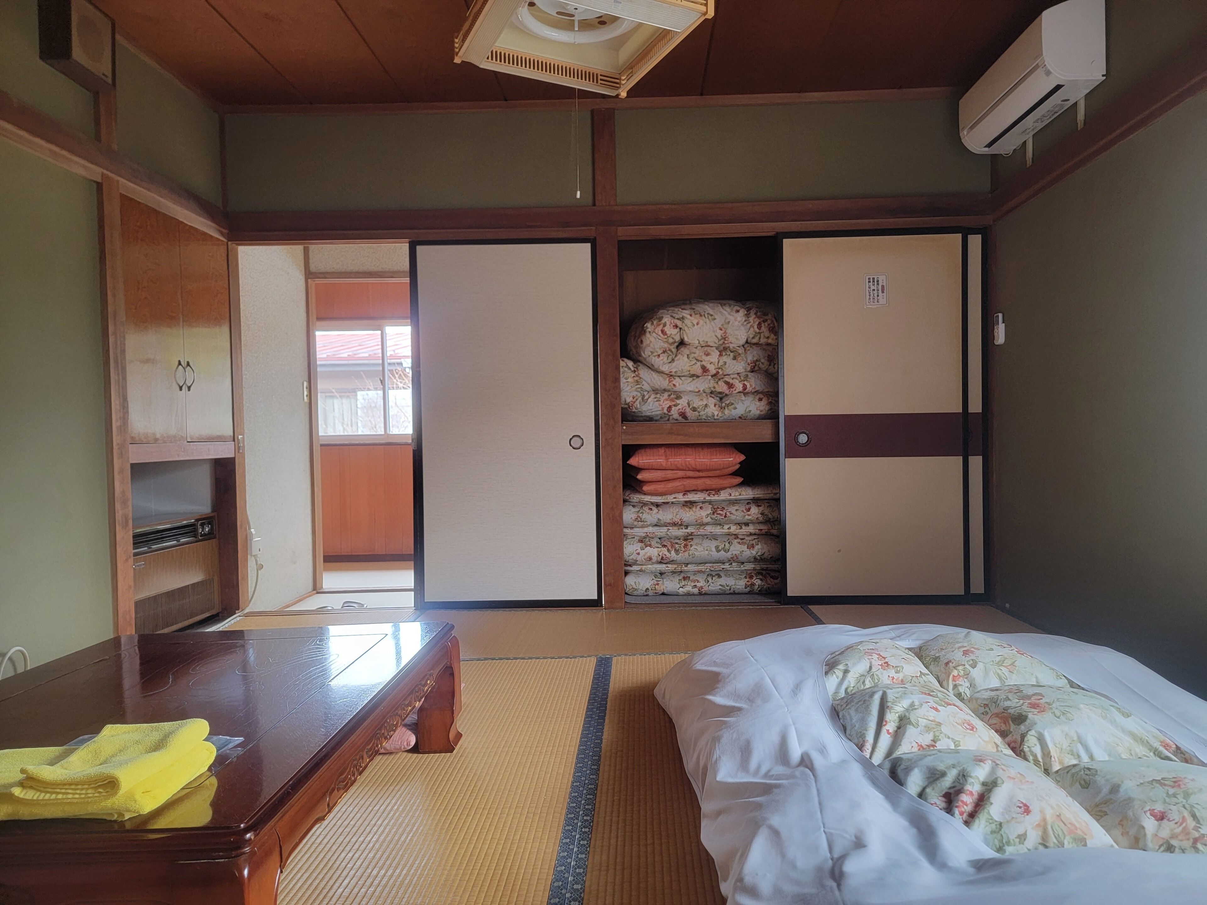 Una habitación tradicional japonesa con suelo de tatami, una cama futón, una mesa de madera, un armario y un mueble para guardar futones.
