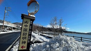 A snow-covered landscape with a signpost featuring Japanese characters, a road, and a body of water in the background.