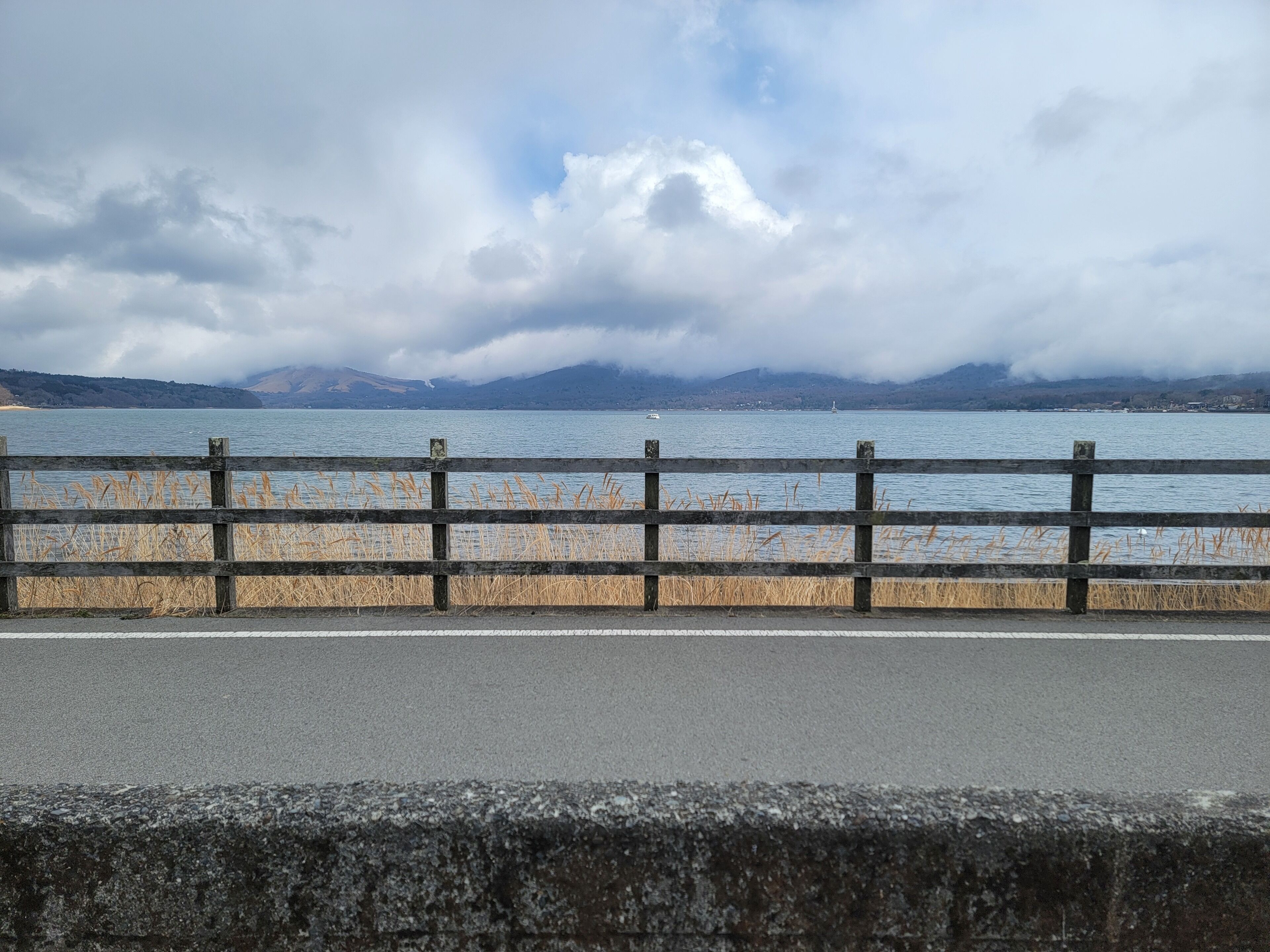 A view of a lake with mountains in the background, a wooden fence in the foreground, and a cloudy sky.
