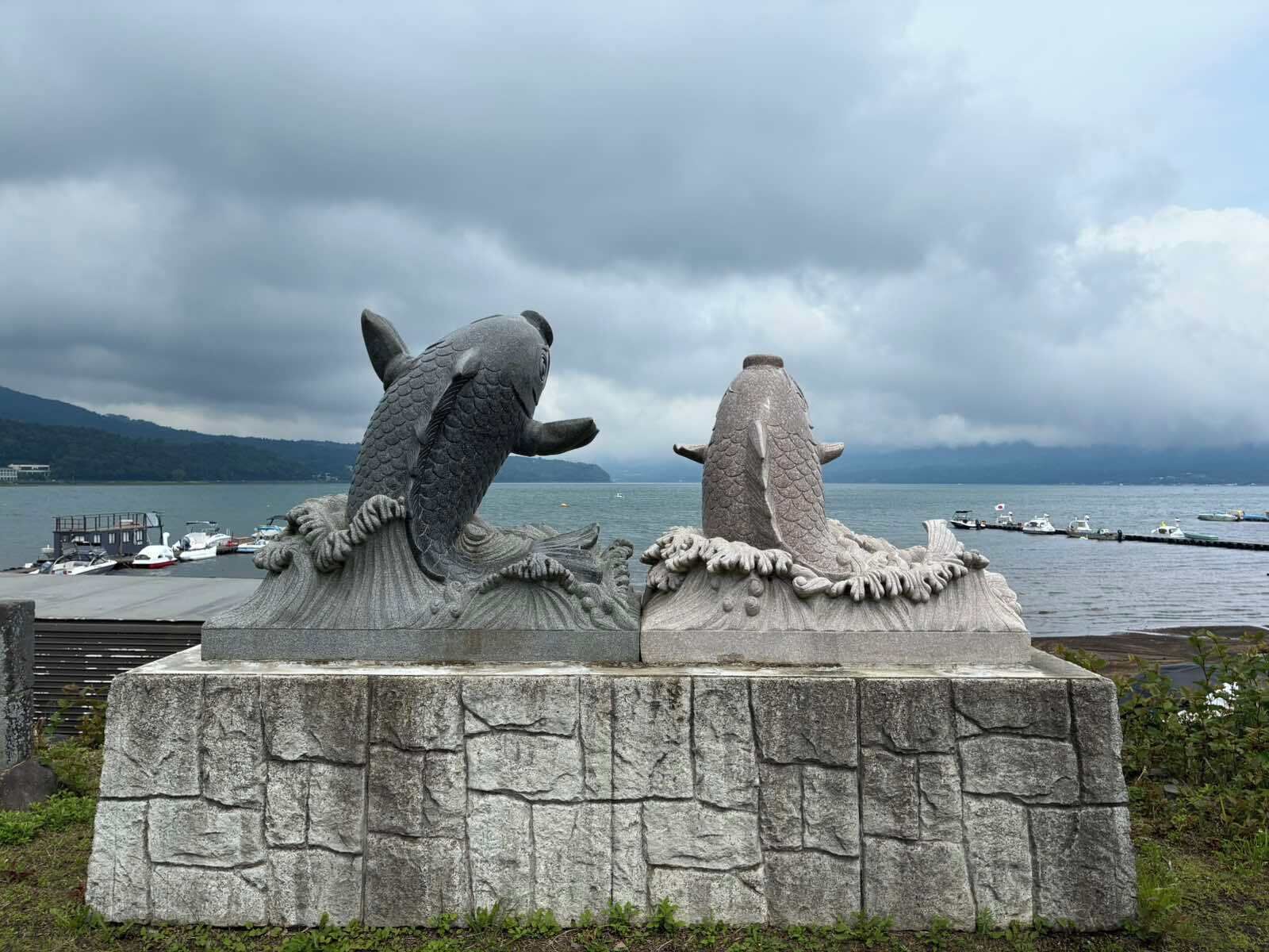 Eine Steinskulptur von zwei Fischen auf einem Sockel am Wasser.