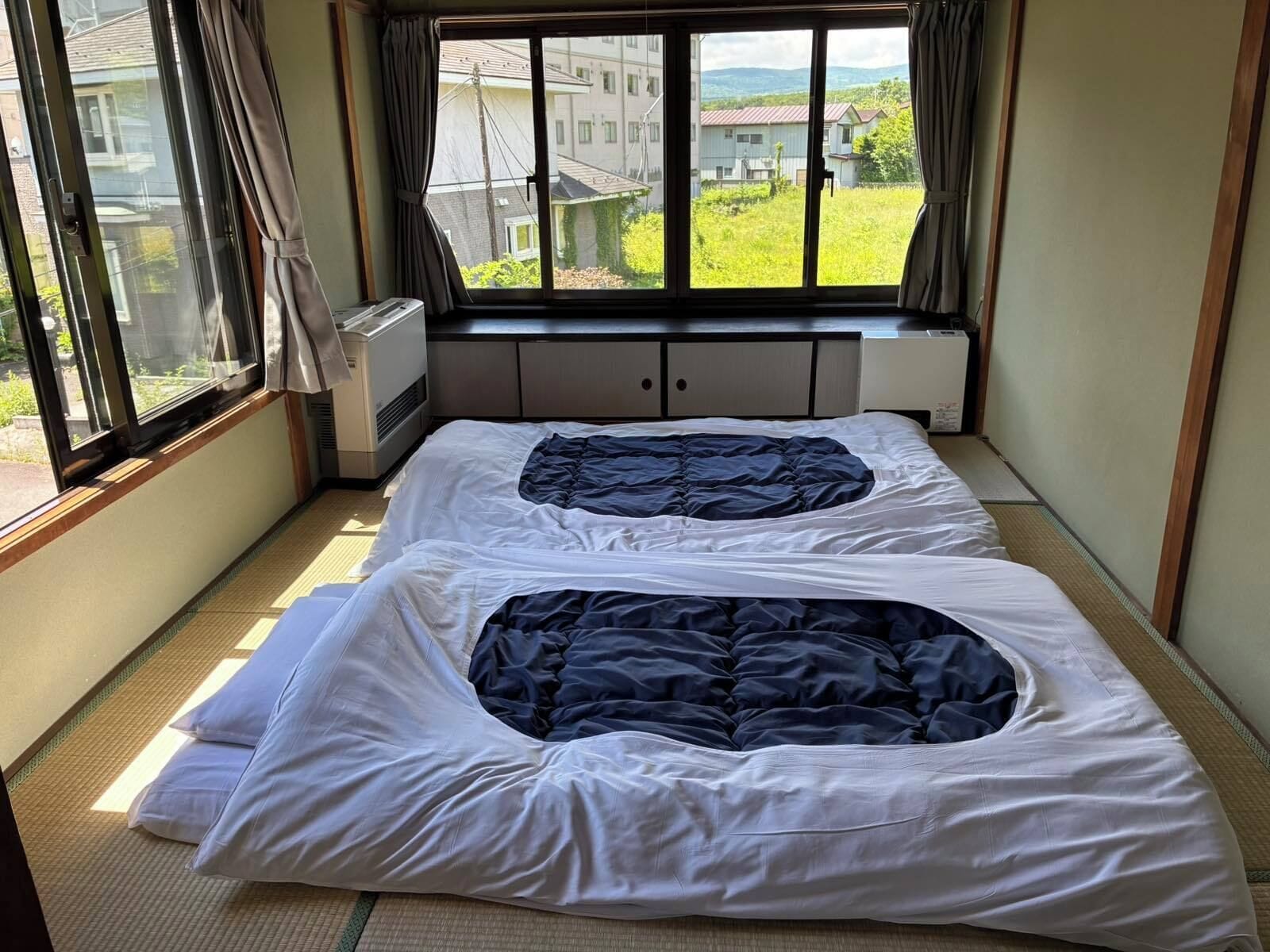 A room with two beds, a window with curtains, a radiator, and a view of buildings and greenery outside.