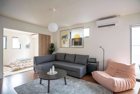 A modern living room with a grey sectional sofa, a round coffee table, a pink pouf, and a large window.