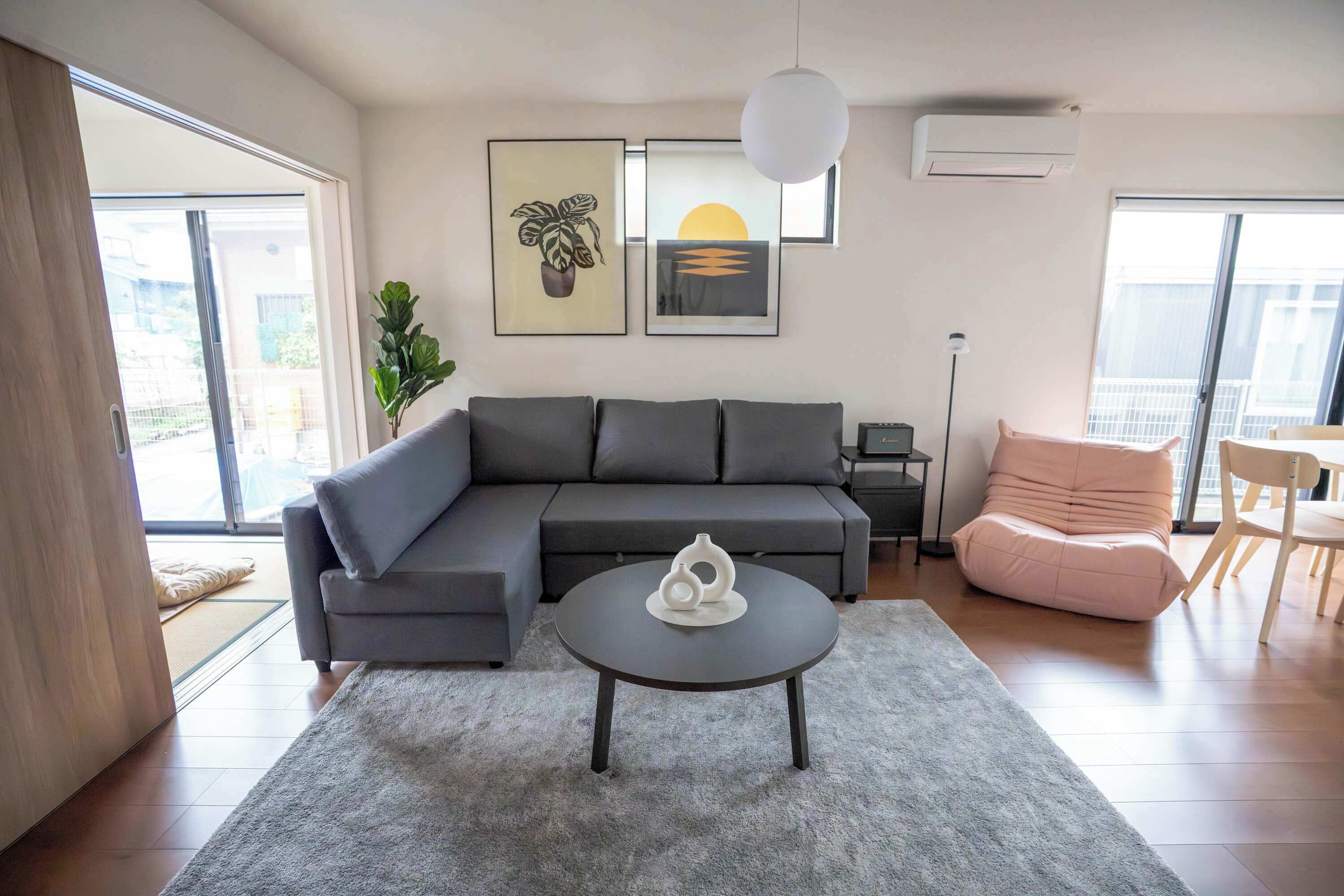 A modern living room with a grey sofa, a round coffee table, and a pink chair. There are two framed pictures on the wall, a potted plant, and a ceiling light.