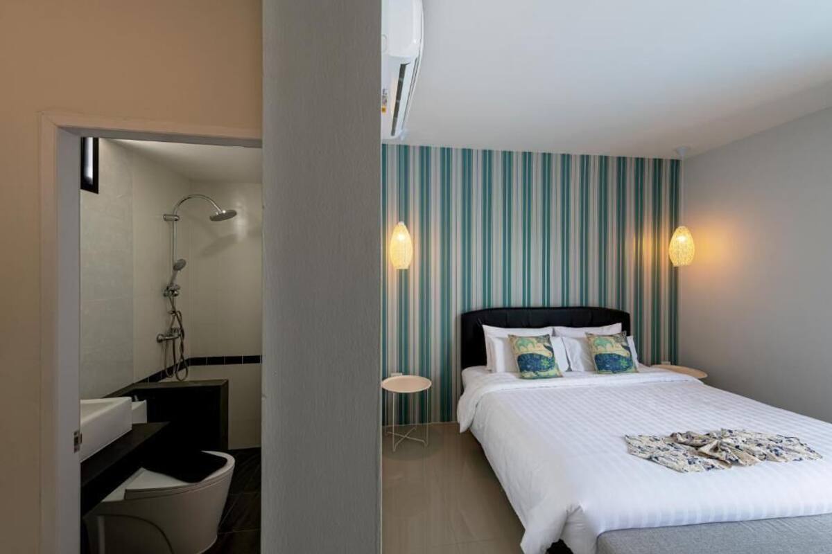 Habitación de hotel con cama, una mesita pequeña redonda, mesita de noche, ducha y pared con rayas verticales.
