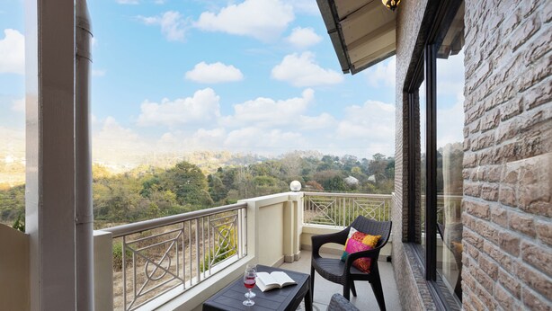 Un balcon donnant sur un paysage vallonné, une table avec un livre et un verre de vin rouge, et deux chaises en osier.