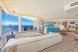A modern living room with a large window, a sofa, a dining area, and a view of the sea. - Celebrate Honeymoon, Anniversary or Birthday...Panoramic Ocean View! (Wailuku)