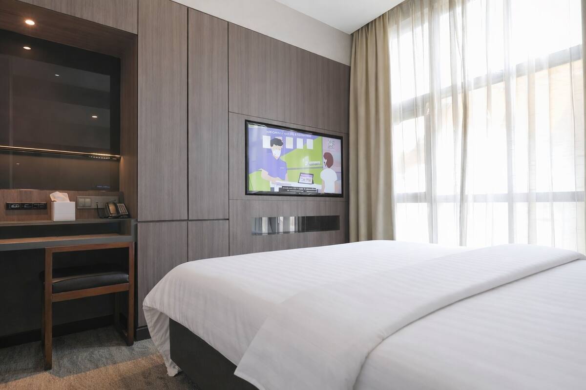 Una habitación de hotel con cama, escritorio, televisor montado en la pared y ventana con cortinas.