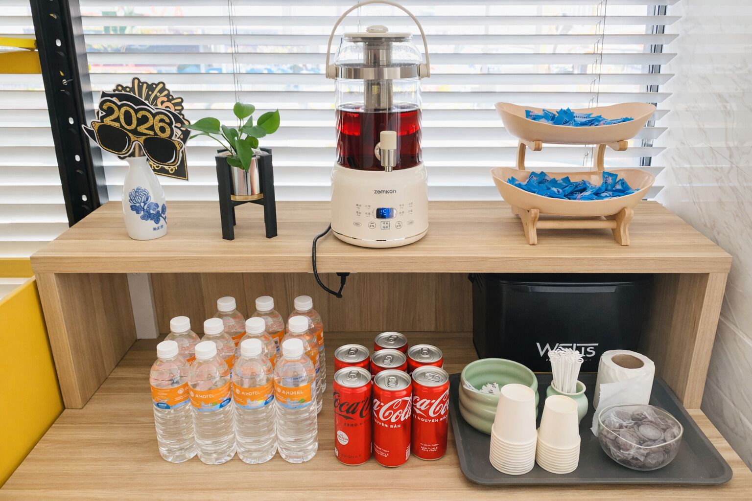Disposición de la nevera del hotel con agua embotellada, latas de Coca-Cola, un fabricante de té y un bol de hielo.